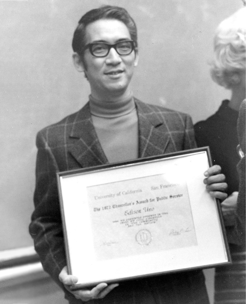 A black and white photo of Edison Uno holding his framed 1972 UCSF Chancellor’s Award for Public Service.